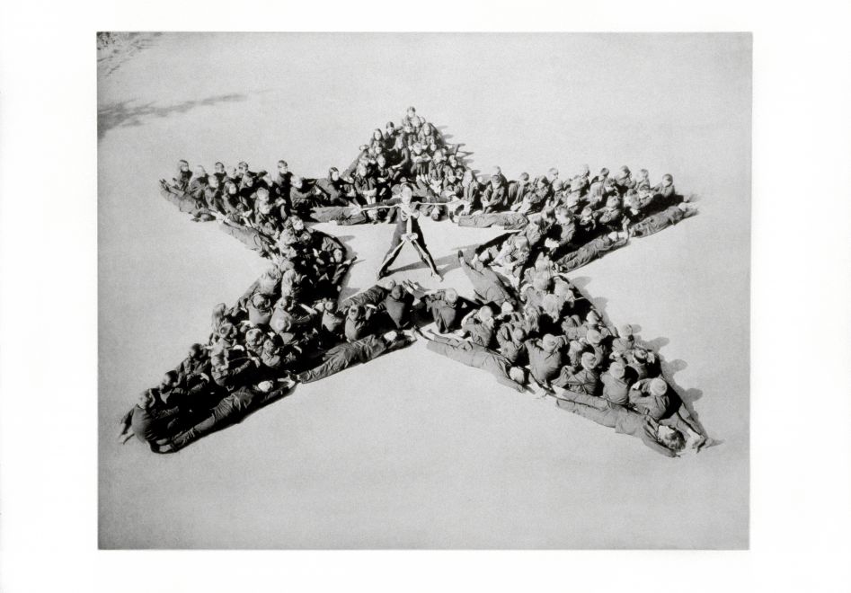 Fotogravüre von der Künstlerin Marina Abramovic. Schwarz-Weiß-Foto von Dutzenden von als Soldaten verkleideten Kindern, die sich in Formation hinlegen, um einen großen fünfzackigen Stern auf dem Boden zu bilden, von oben gesehen. Marina Abramovic steht mit ausgestreckten Armen und Beinen in der Mitte des Sterns und trägt schwarze Kleidung, auf deren Vorderseite ein Skelett angebracht ist.