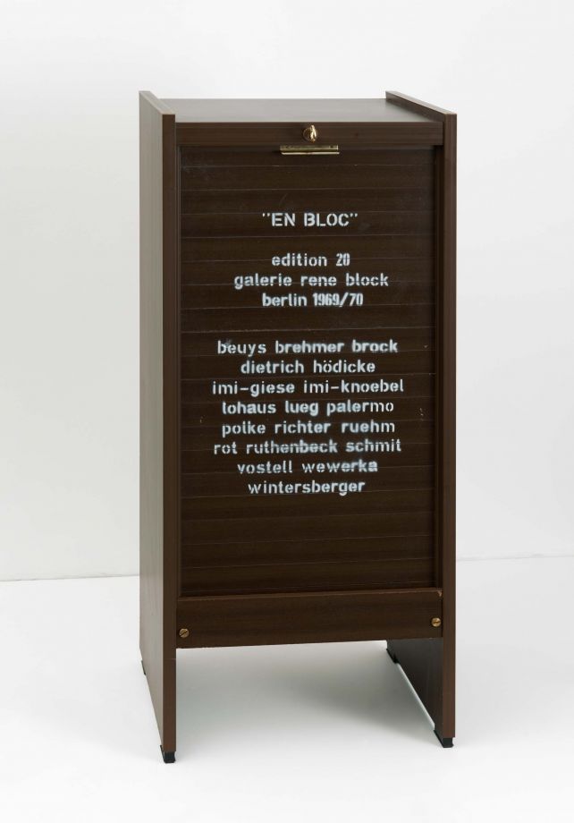 Edition En Bloc. Ein dunkelbrauner, geschlossener Rolladenschrank vor einer weißen Wand. Auf der Vorderseite ist mit weißer Farbe gestempelt: EN BLOC. edition 20. galerie rene block. berlin 1969/70. beuys brehmer brock diedrich hödicke imi-giese imi-knoebel lohaus lueg palermo polke richter ruehm rot ruthenbeck schmit vostell wewerka wintersberger