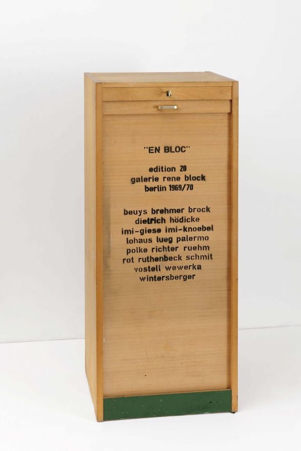 Edition En Bloc. Ein hellbrauner, geschlossener Rolladenschrank vor einer weißen Wand. Auf der Vorderseite ist mit schwarzer Farbe gestempelt: EN BLOC. edition 20. galerie rene block. berlin 1969/70. beuys brehmer brock diedrich hödicke imi-giese imi-knoebel lohaus lueg palermo polke richter ruehm rot ruthenbeck schmit vostell wewerka wintersberger